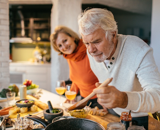 Older man preparing a meal in his kitchen while a family member looks on